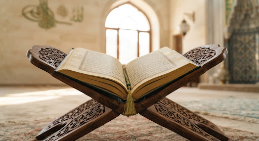 Close up photography of an open Holy Quran with Arabic calligraphy lying on a carved wooden rehal bookstand in a peaceful sunlit mosque setting with subtle green and gold highlights